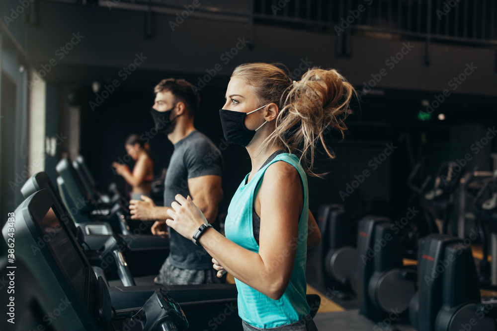 Young fit woman and man running on treadmill in modern fitness gym. They keeping distance and wearing protective face masks. Coronavirus world pandemic and sport theme.