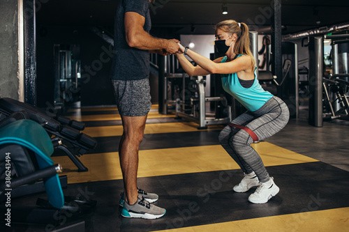 Young fit and attractive woman at body workout in modern gym together with her personal fitness instructor or coach. They wearing protective face masks. Coronavirus world pandemic and sport theme.