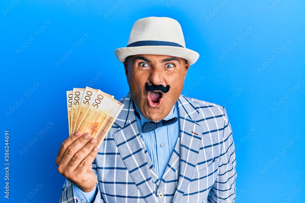 Mature middle east man with mustache wearing elegant vintage style holding norwegian krone scared and amazed with open mouth for surprise, disbelief face Mature middle east man with mustache wearing elegant vintage style holding norwegian krone scared and amazed with open mouth for surprise, disbelief face