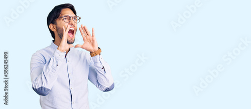 Handsome hispanic man wearing business shirt and glasses shouting angry out loud with hands over mouth