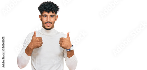 Young arab man wearing casual winter sweater success sign doing positive gesture with hand, thumbs up smiling and happy. cheerful expression and winner gesture.
