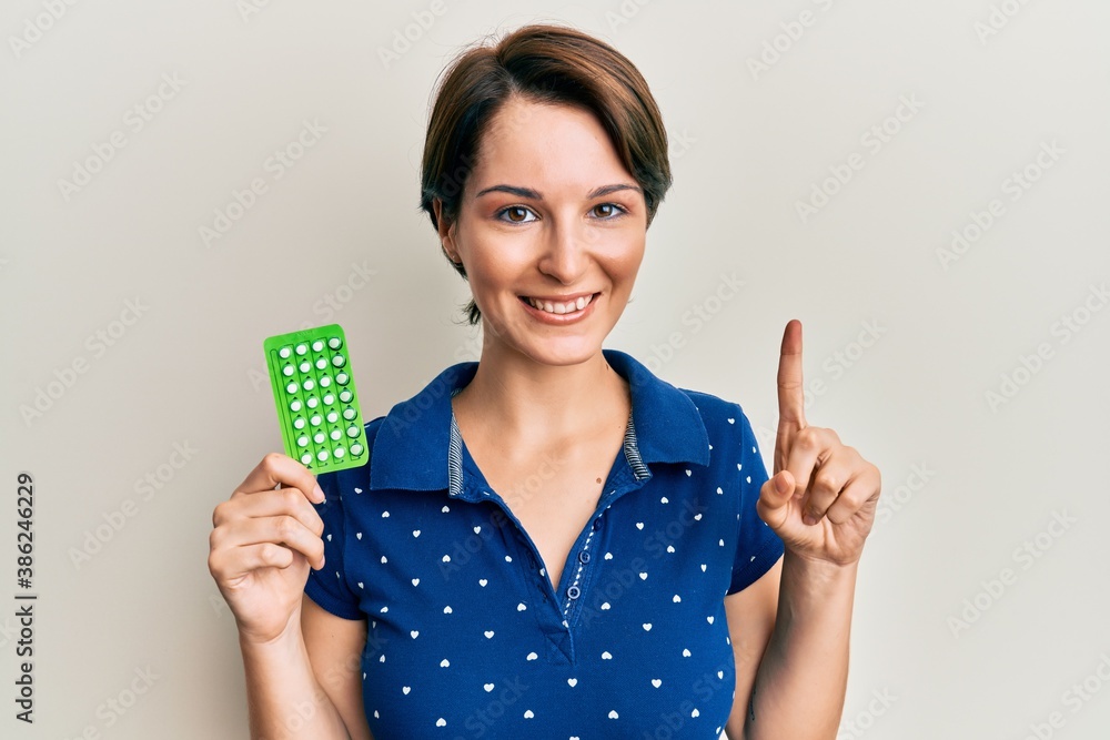 Young brunette woman with short hair holding birth control pills smiling with an idea or question pointing finger with happy face, number one Young brunette woman with short hair holding birth control pills smiling with an idea or question pointing finger with happy face, number one