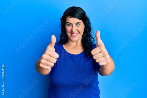 Plus size brunette woman wearing casual blue shirt approving doing positive gesture with hand, thumbs up smiling and happy for success. winner gesture. © Krakenimages.com