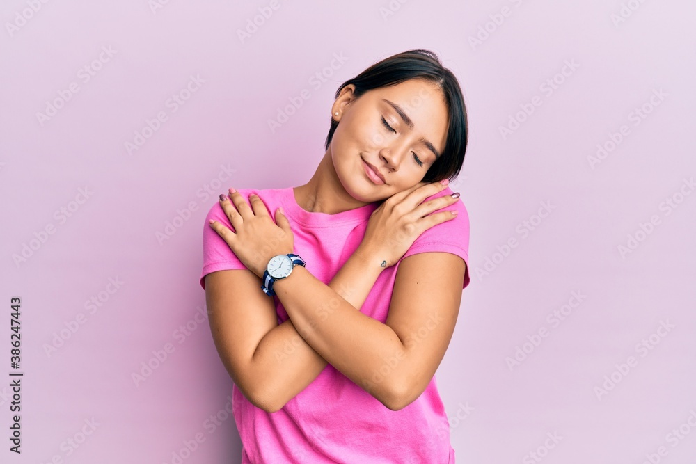 Beautiful young woman with short hair wearing pink tshirt over pink background hugging oneself happy and positive, smiling confident. self love and self care Beautiful young woman with short hair wearing pink tshirt over pink background hugging oneself happy and positive, smiling confident. self love and self care