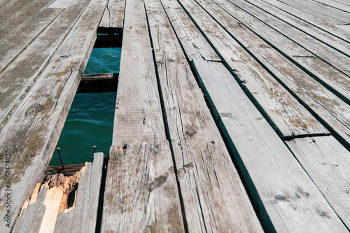An old wooden dock with a broken board and a hole in the floor where the blue surface of the sea is visible