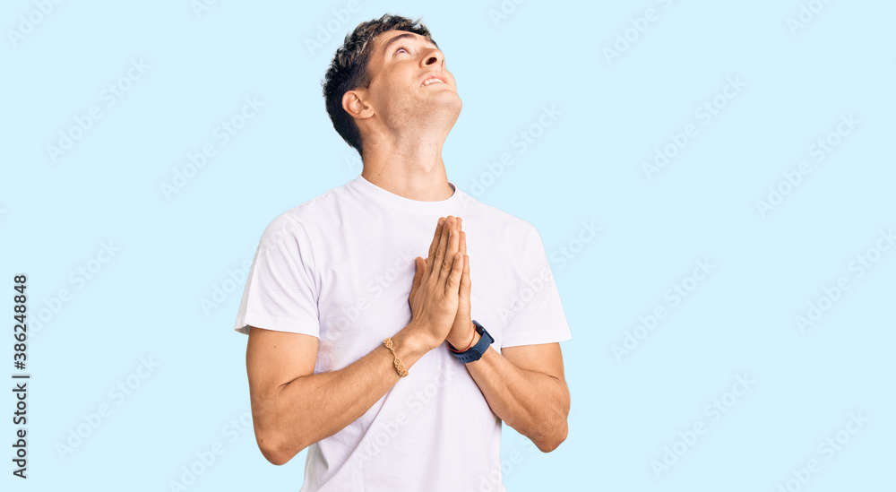 Young handsome man wearing casual white tshirt begging and praying with hands together with hope expression on face very emotional and worried. begging. Young handsome man wearing casual white tshirt begging and praying with hands together with hope expression on face very emotional and worried. begging.