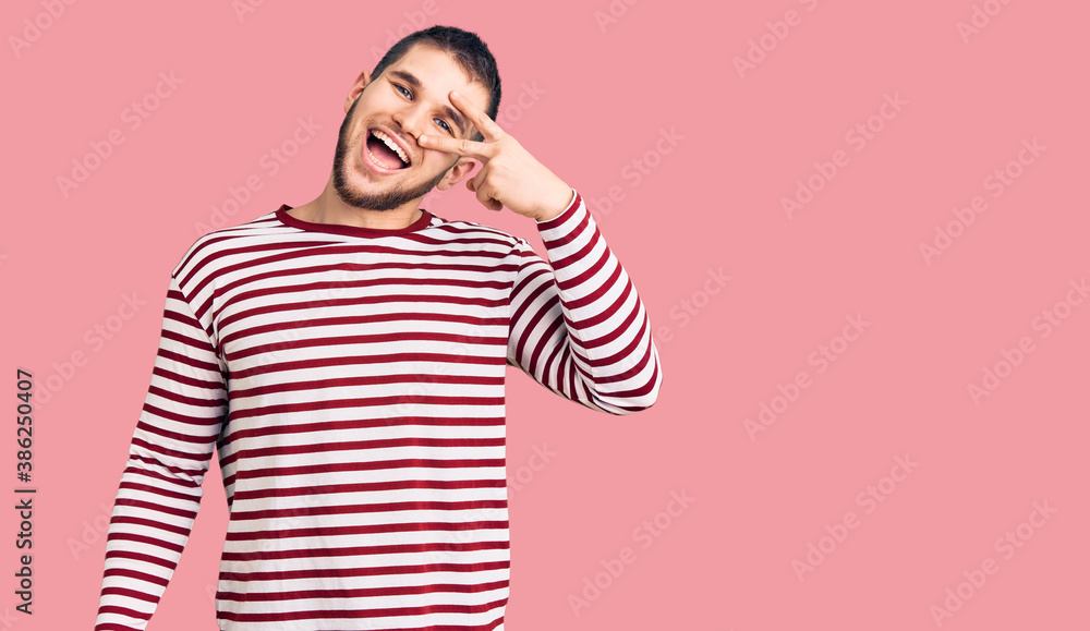 Young handsome man wearing striped sweater doing peace symbol with fingers over face, smiling cheerful showing victory Young handsome man wearing striped sweater doing peace symbol with fingers over face, smiling cheerful showing victory