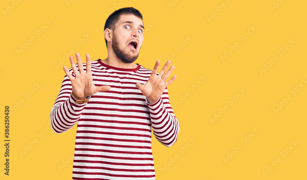 Young handsome man wearing striped sweater afraid and terrified with fear expression stop gesture with hands, shouting in shock. panic concept. Young handsome man wearing striped sweater afraid and terrified with fear expression stop gesture with hands, shouting in shock. panic concept.