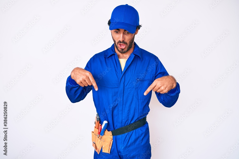 Handsome young man with curly hair and bear weaing handyman uniform pointing down with fingers showing advertisement, surprised face and open mouth