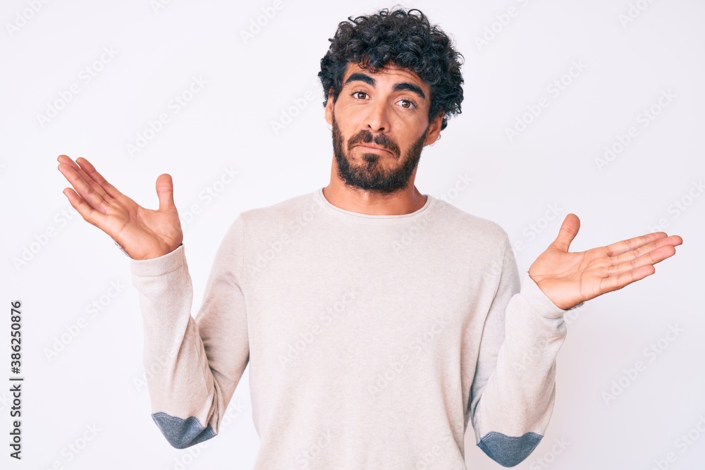 Handsome young man with curly hair and bear wearing casual winter sweater clueless and confused with open arms, no idea and doubtful face. Handsome young man with curly hair and bear wearing casual winter sweater clueless and confused with open arms, no idea and doubtful face.