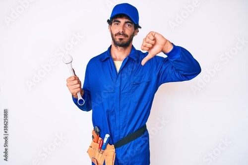 Handsome young man with curly hair and bear wearing builder jumpsuit uniform and holding wrench with angry face, negative sign showing dislike with thumbs down, rejection concept