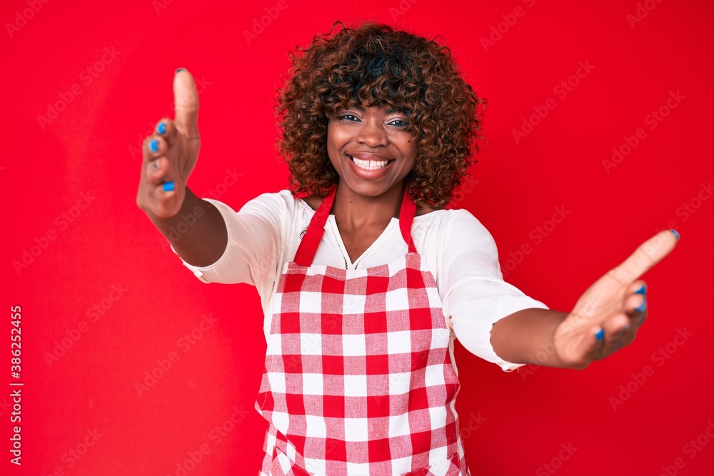 Young african american woman wearing apron looking at the camera smiling with open arms for hug. cheerful expression embracing happiness.