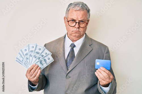 Senior grey-haired man wearing business suit holding credit car and dollars skeptic and nervous, frowning upset because of problem. negative person.