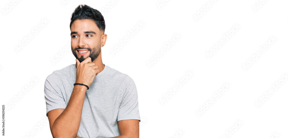 Fototapeta premium Young man with beard wearing casual grey tshirt with hand on chin thinking about question, pensive expression. smiling with thoughtful face. doubt concept.