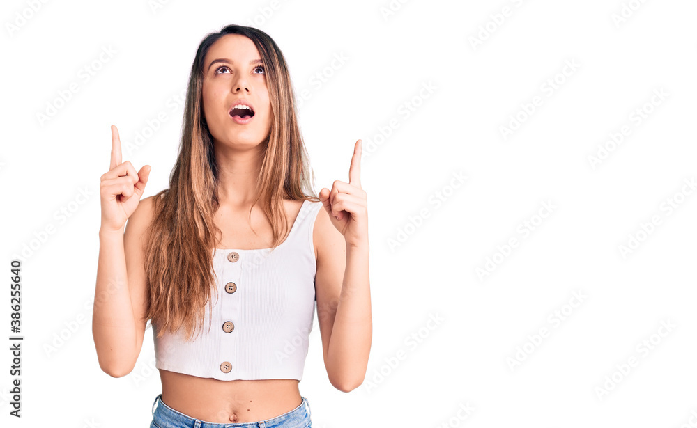 Young beautiful girl wearing casual sleeveless t shirt amazed and surprised looking up and pointing with fingers and raised arms. Young beautiful girl wearing casual sleeveless t shirt amazed and surprised looking up and pointing with fingers and raised arms.