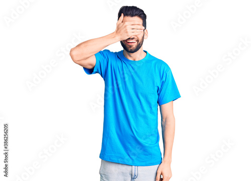 Young handsome man with beard wearing casual t-shirt smiling and laughing with hand on face covering eyes for surprise. blind concept.