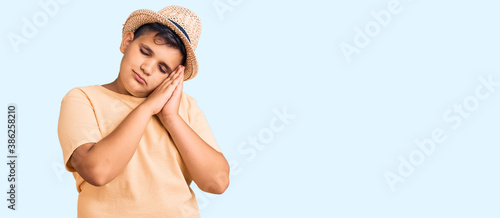 Little boy kid wearing summer hat and hawaiian swimsuit sleeping tired dreaming and posing with hands together while smiling with closed eyes.