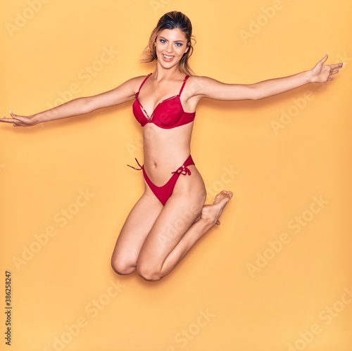 Young beautiful girl on vacation wearing bikini smiling happy. Jumping with smile on face and arms opened over isolated yellow background © Krakenimages.com