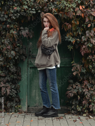 Full length portrait young redhead woman with autumn leaf in hand and fall red and yellow ivy background enjoying fall day