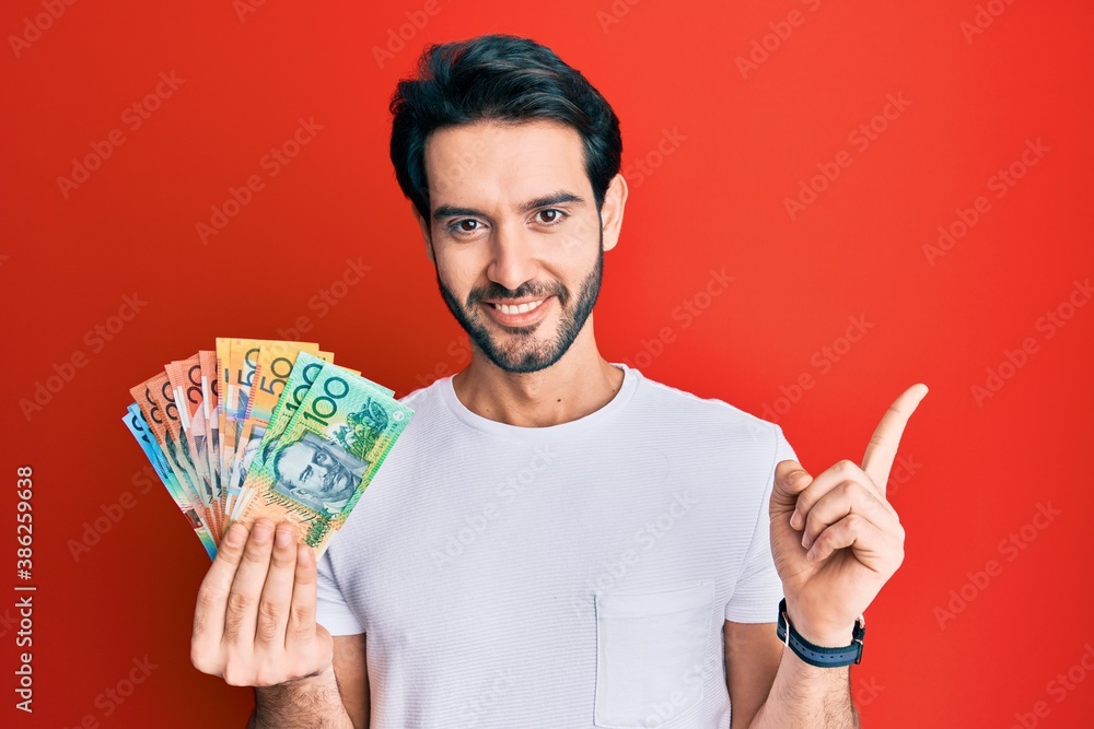 Young hispanic man holding australian dollars smiling happy pointing with hand and finger to the side Young hispanic man holding australian dollars smiling happy pointing with hand and finger to the side