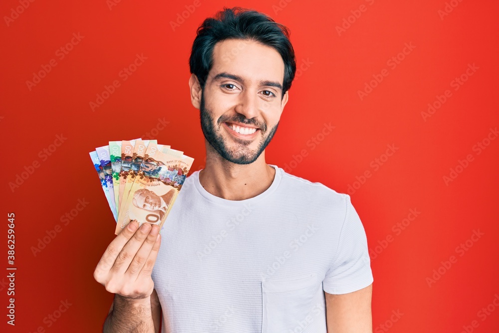 Young hispanic man holding canadian dollars looking positive and happy standing and smiling with a confident smile showing teeth Young hispanic man holding canadian dollars looking positive and happy standing and smiling with a confident smile showing teeth