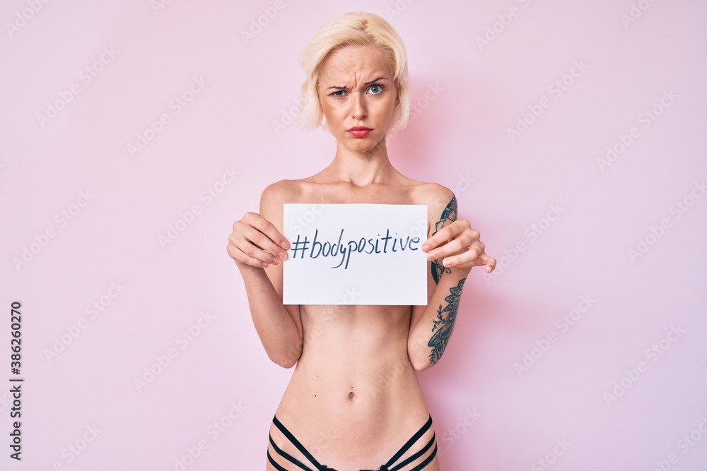 Young blonde woman with tattoo standing shirtless holding banner with body positive message skeptic and nervous, frowning upset because of problem. negative person. Young blonde woman with tattoo standing shirtless holding banner with body positive message skeptic and nervous, frowning upset because of problem. negative person.