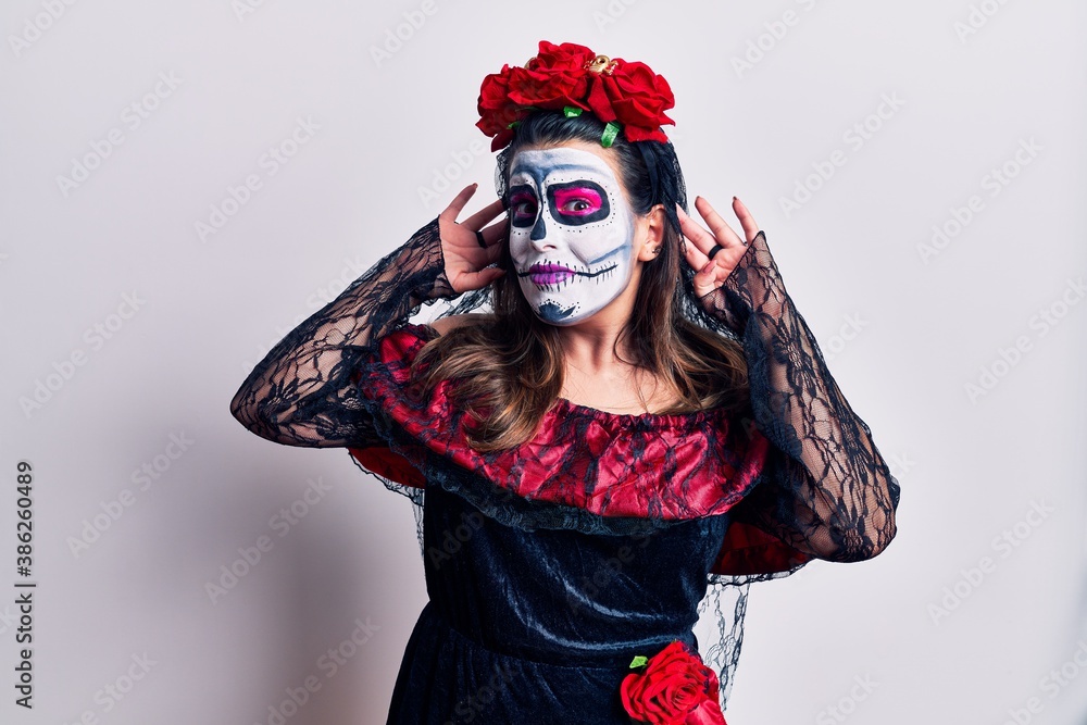 Young woman wearing day of the dead costume over white trying to hear both hands on ear gesture, curious for gossip. hearing problem, deaf