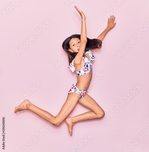Adorable hispanic child girl on vacation wearing bikini smiling happy. Jumping with smile on face over isolated pink background