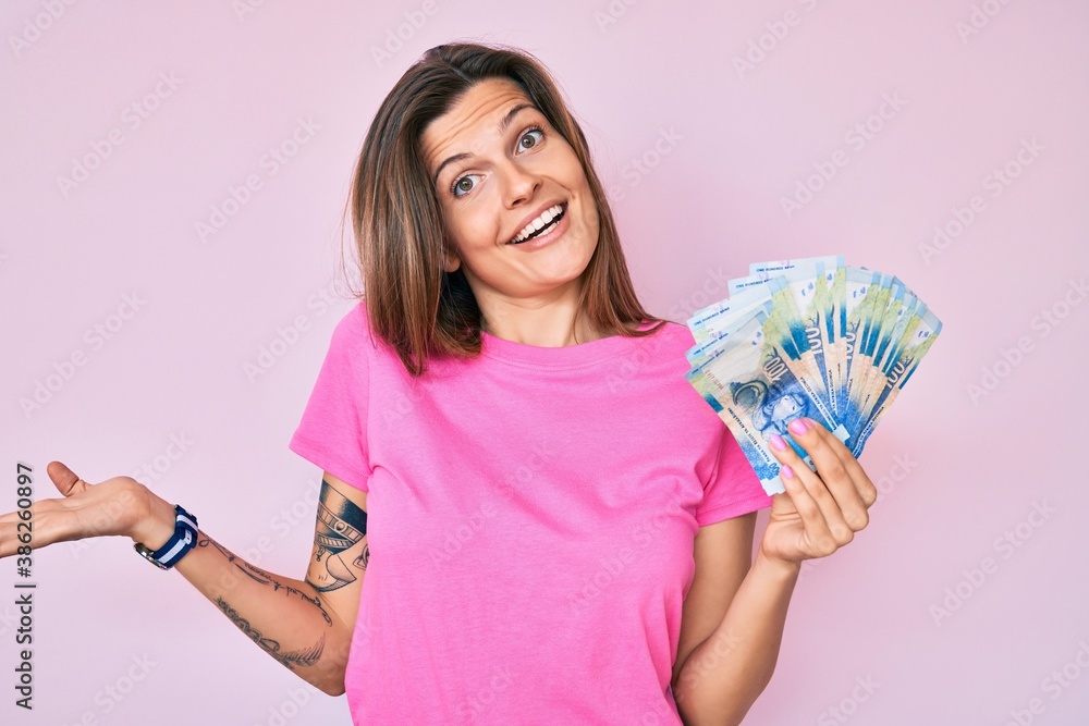 Beautiful caucasian woman holding south african rand banknotes celebrating achievement with happy smile and winner expression with raised hand Beautiful caucasian woman holding south african rand banknotes celebrating achievement with happy smile and winner expression with raised hand