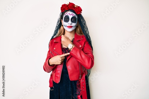 Woman wearing day of the dead costume over white in hurry pointing to watch time, impatience, looking at the camera with relaxed expression