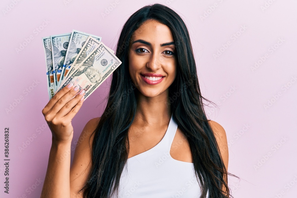 Beautiful hispanic woman holding dollars looking positive and happy standing and smiling with a confident smile showing teeth Beautiful hispanic woman holding dollars looking positive and happy standing and smiling with a confident smile showing teeth