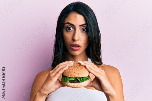Beautiful hispanic woman eating a tasty classic burger in shock face  looking skeptical and sarcastic  surprised with open mouth