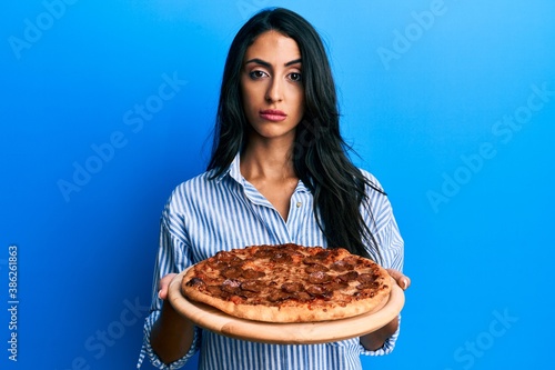 Beautiful hispanic woman holding tasty pepperoni pizza skeptic and nervous  frowning upset because of problem. negative person.