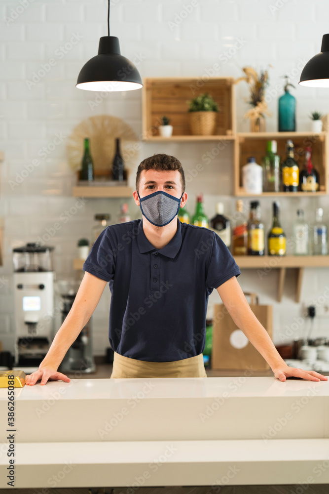 portrait of a professional waiter with mask posing behind the bar of a restaurant during coronavirus pandemic