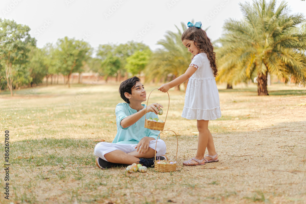 Latin brothers celebrating Easter. Boy sitting on the grass holding a easter egg and curly girl with a hamper on a sunny day