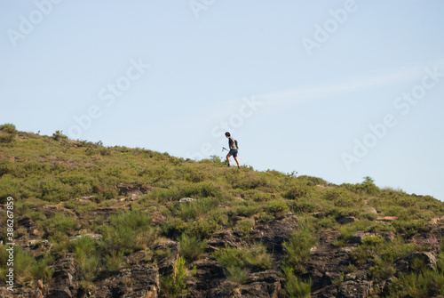 Pessoa, homem a caminhar na montanha a subir uma encosta inclinada com equipamento ás costas num dia de sol, trail running