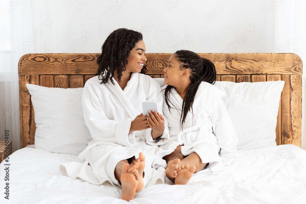 African Mom And Daughter Using Smartphone While Relaxing On Bed In Bathrobes African Mom And Daughter Using Smartphone While Relaxing On Bed In Bathrobes