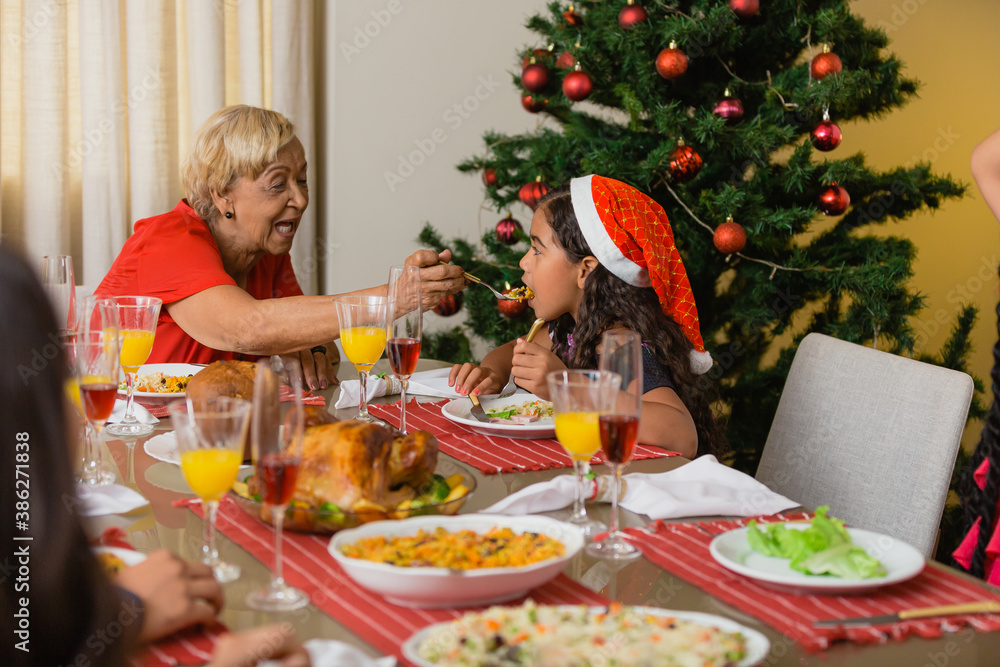 Summer Christmas dinner in Brazil. Real Brazilian family having fun at the Latin American Christmas party. elderly woman giving dinner in the mouth of a curly haired child Summer Christmas dinner in Brazil. Real Brazilian family having fun at the Latin American Christmas party. elderly woman giving dinner in the mouth of a curly haired child