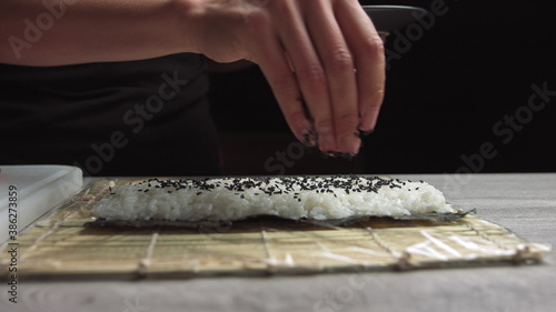 Chef's hands sprinkle sesame seeds on the inside out sushi. Japanese chef in black gloves at work preparing sushi roll with salmon and avocado.