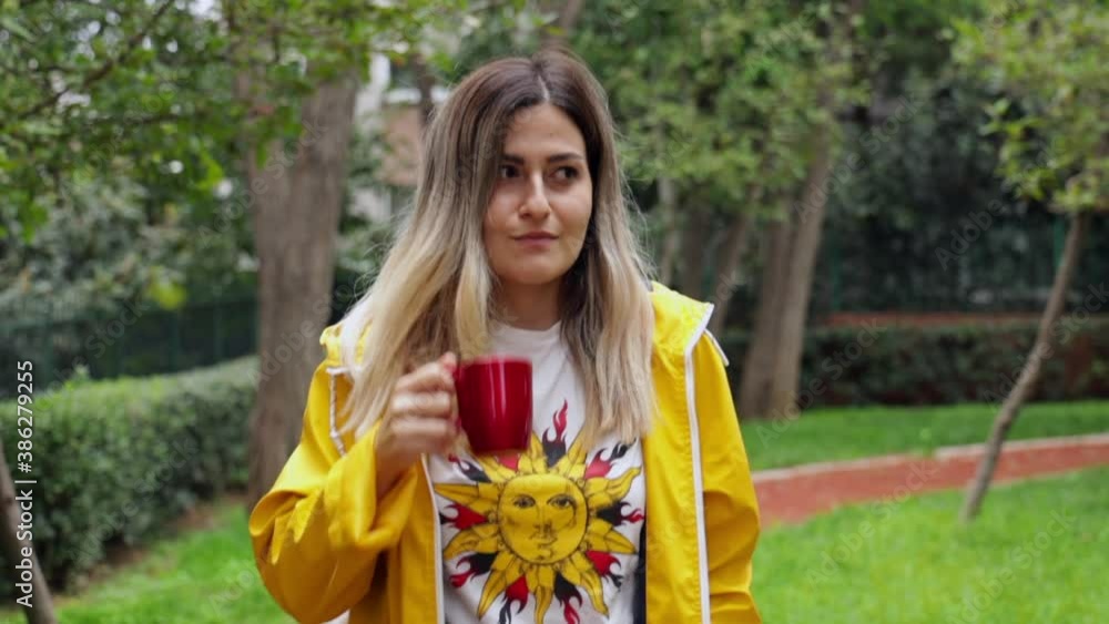 Young attractive girl with yellow raincoat walking in the park, drinking coffee from red traveling mug. Girl in style, during a cloudy autumn day surrounded by green trees and grasses Young attractive girl with yellow raincoat walking in the park, drinking coffee from red traveling mug. Girl in style, during a cloudy autumn day surrounded by green trees and grasses