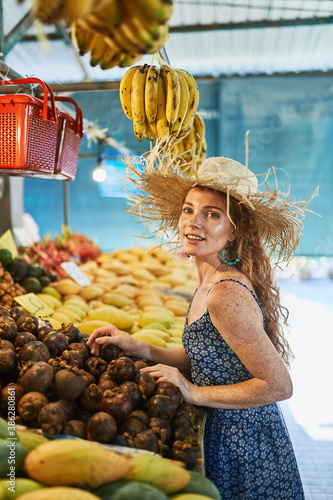Young redhead smiling girl with freckles in a blue dress and a straw hat chooses fruits at the market in Thailand © Nikolay