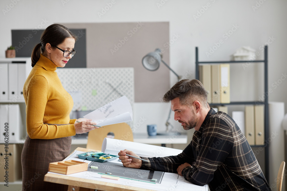 Side view portrait of two adult architects discussing blueprints while working together in office