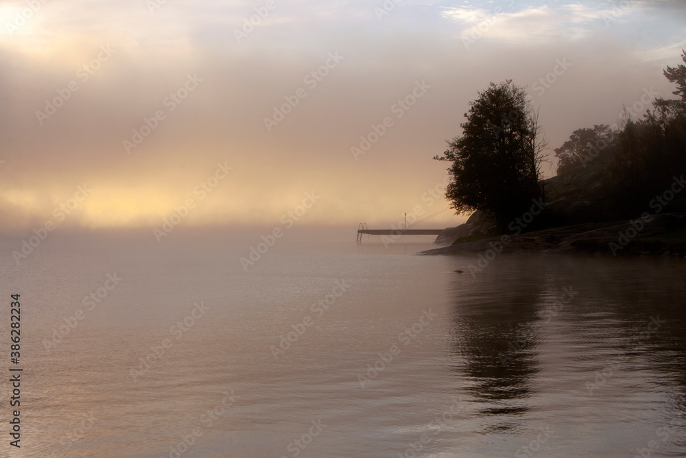 Sunrise and fog in southern Norway - silhouette of tree and small jetty with calm sea