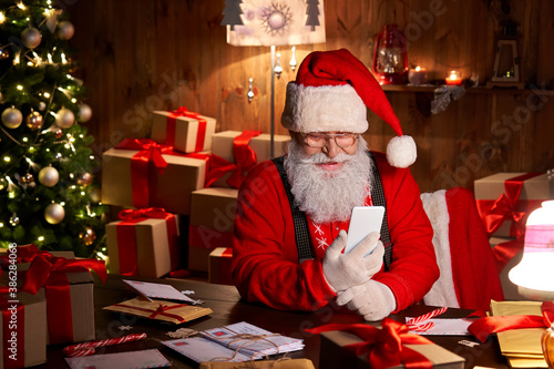 Happy Santa Claus holding smartphone holding cell phone using mobile app for social distance communication, watching videos or video calling in virtual online chat sit at home table late on xmas eve. photo