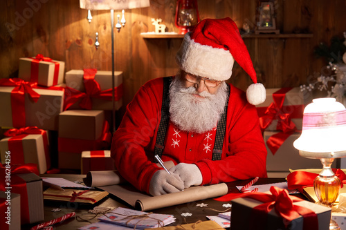 Happy old kind bearded Santa Claus wearing hat, glasses, writing on wish list, working on Christmas eve sitting at cozy home workshop table late with presents, tree and candles preparing for holidays. photo