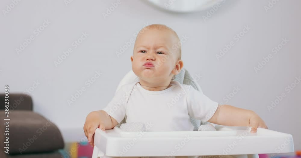 Portrait of young baby in white high baby chair who makes funny faces and grimaces. White walls on background. Portrait of young baby in white high baby chair who makes funny faces and grimaces. White walls on background.