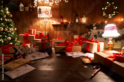 Santa Claus workshop home wooden decorated table with Merry Christmas tree, decor, wrapped gifts presents boxes on holiday eve in cozy home interior late in night with lamp light on xmas background. photo