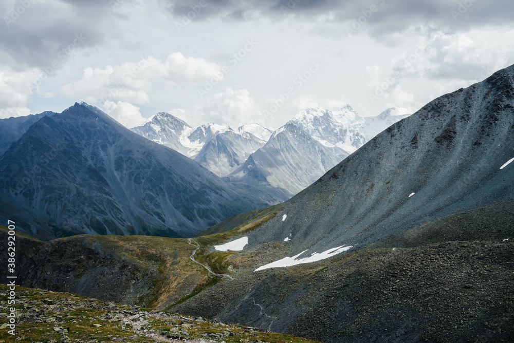 Trail through stony hills to beautiful huge glacier mountains under gray cloudy sky. Awesome dramatic alpine landscape with snow mountains and glaciers. Atmospheric highland scenery. Bad rainy weather