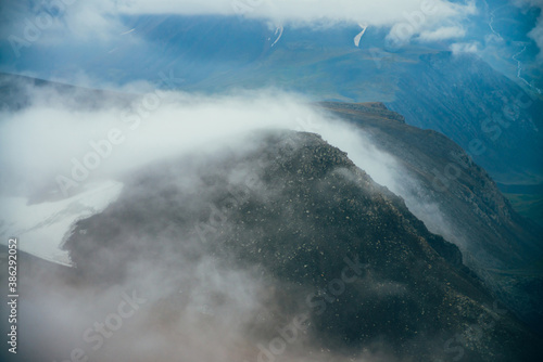 Atmospheric minimalist alpine landscape to giant low cloud among rocky mountain with snow. Low clouds among rockies in valley. Wonderful tranquil highland scenery. Flying over mountains above clouds.