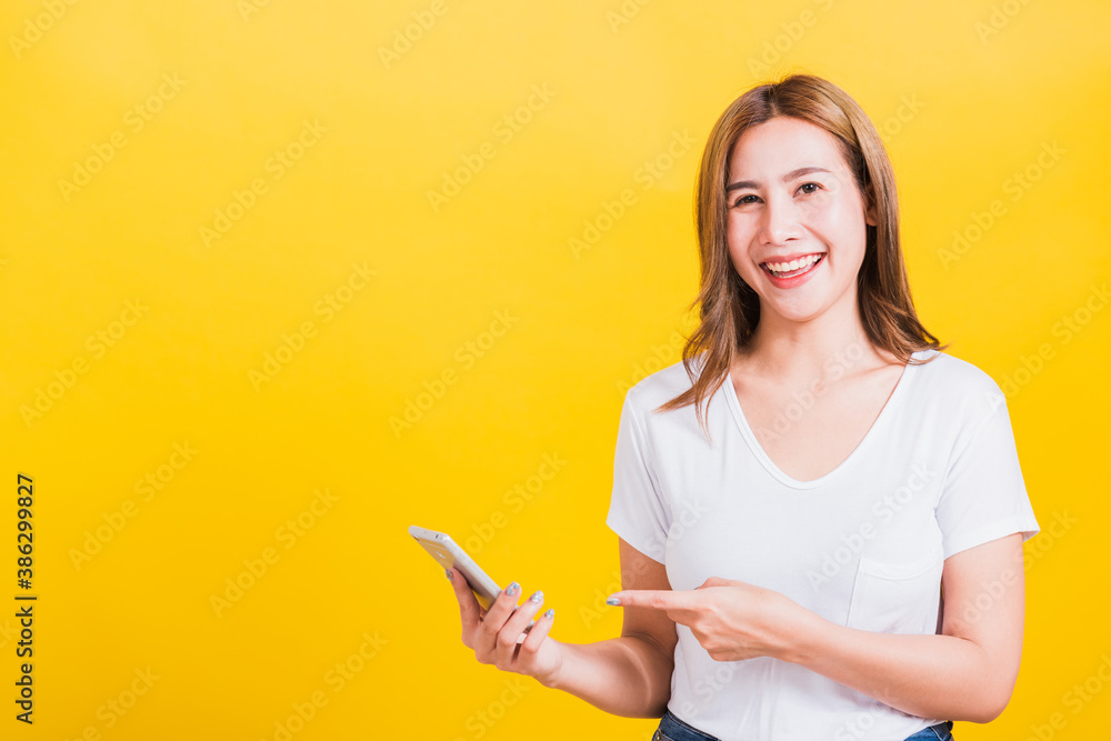 Asian Thai portrait happy beautiful young woman smile standing wear t-shirt making finger pointing on smartphone blank screen looking to camera isolated, studio shot yellow background with copy space